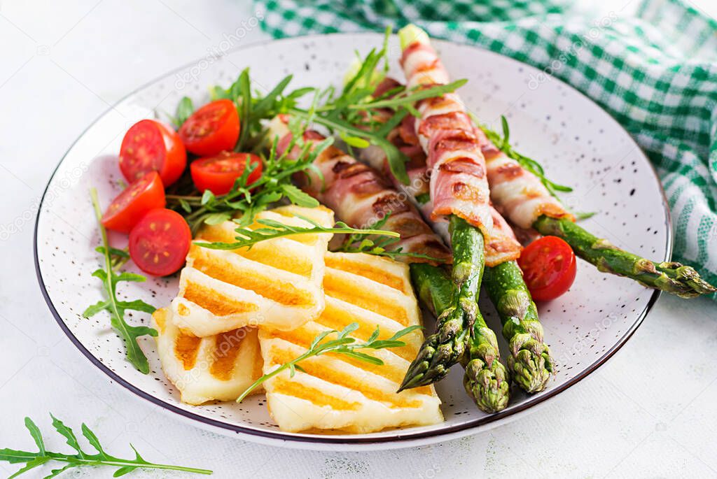 Grilled halloumi cheese salad with tomatoes and asparagus in strips of bacon on plate on light background. Healthy food.