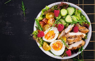 Izgara tavuk eti ve taze sebze salatası. Domates, salatalık, yumurta, marul ve ahududu. Ketogenik diyet. Buda çanağı koyu arkaplanda. Üst manzara, düz uzanma