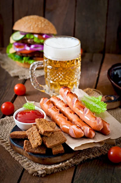 Grote hamburger, gegrilde worstjes met croutons en bier — Stockfoto