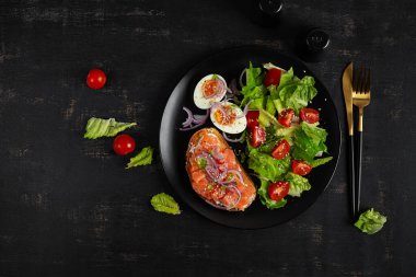 Lezzetli sağlıklı kahvaltı - haşlanmış yumurta, somon balığı ve yeşil salata. Koyu arkaplanda dengeli yemek tabağı, üst görünüm, kopyalama alanı.