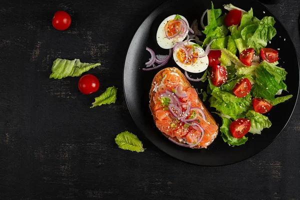 Lezzetli sağlıklı kahvaltı - haşlanmış yumurta, somon balığı ve yeşil salata. Koyu arkaplanda dengeli yemek tabağı, üst görünüm, kopyalama alanı.