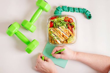 Kızarmış tavuklu sağlıklı beslenme çantası, mercimek, taze sebze salatası ve masanın üzerinde dambıllarla deftere yazan kadın. Üst görünüm