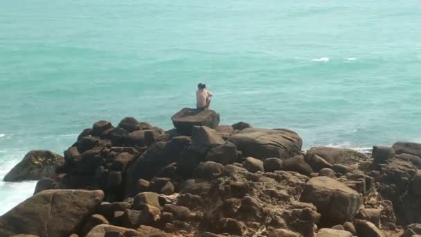 Man zit op een reusachtig rotsblok op een strand en kijken naar de zee — Stockvideo