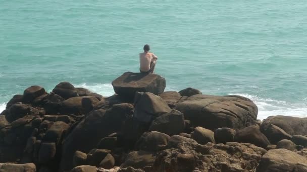 Hombre sentado en una enorme roca en una playa y mirando al mar — Vídeo de stock