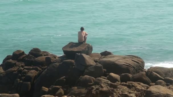 Man sitter på ett stort stenblock på stranden och titta på havet — Stockvideo