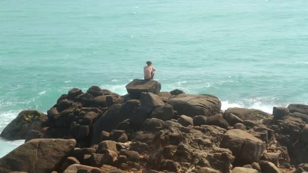 Man sitter på ett stort stenblock på stranden och titta på havet — Stockvideo