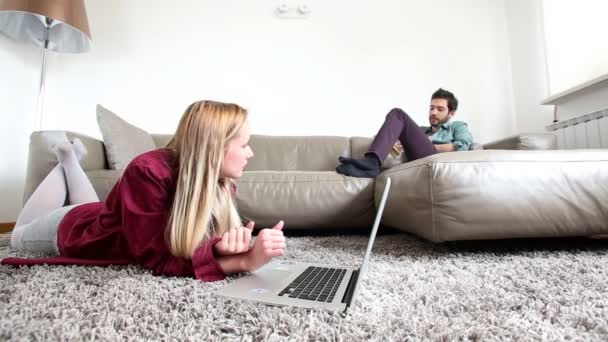 Mujer en la alfombra mirando portátil con el hombre en el sofá — Vídeos de Stock