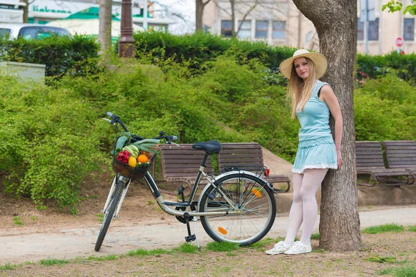 Attractive blonde girl with straw hat standing in the park and posing next to bike with basket full of groceries. — Stock Photo, Image