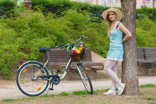 Attractive blonde girl with straw hat standing in the park and posing next to bike with basket full of groceries. — Stock Photo, Image