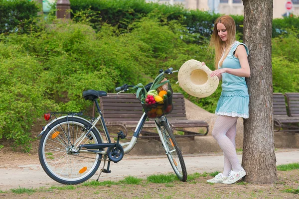 Attraktive blonde Mädchen mit Strohhut steht im Park und posiert neben Fahrrad mit Korb voller Lebensmittel. — Stockfoto