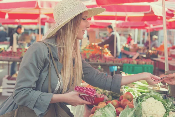 Atractiva mujer rubia con sombrero de paja pagando verduras en el mercado . —  Fotos de Stock