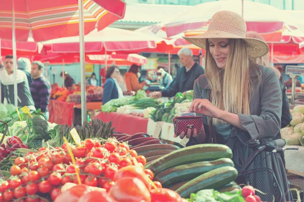 Belle femme blonde avec chapeau de paille payer des légumes sur le marché . — Photo