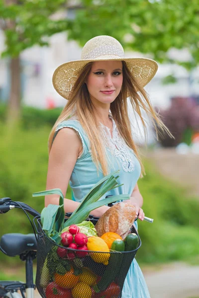 Attraente ragazza bionda con cappello di paglia in piedi nel parco e in posa accanto alla bici con cesto pieno di generi alimentari . — Foto Stock