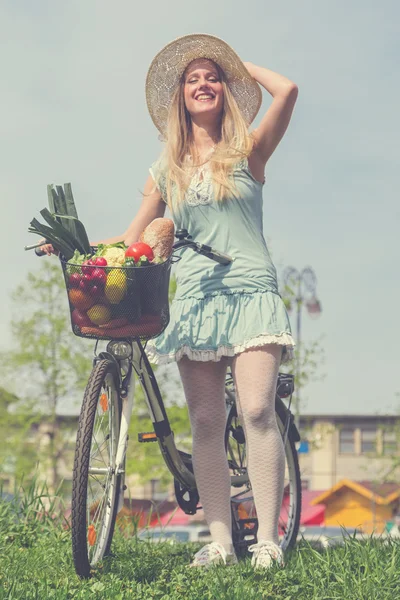 Mulher loira atraente com chapéu de palha posando ao lado de bicicleta com cesta cheia de mantimentos . — Fotografia de Stock