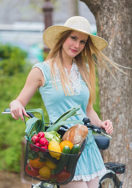 Attractive blonde girl with straw hat standing in the park and posing next to bike with basket full of groceries. — Stock Photo, Image