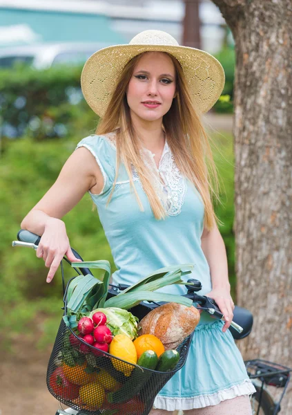 Menina loira atraente com chapéu de palha em pé no parque e posando ao lado de bicicleta com cesta cheia de compras . — Fotografia de Stock