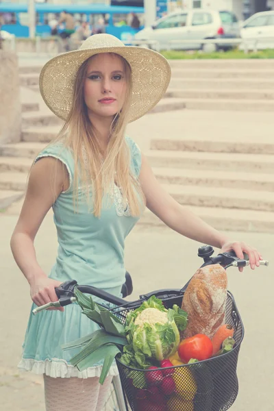 Attraktive blonde Frau mit Strohhut posiert neben Fahrrad mit Korb voller Lebensmittel. — Stockfoto