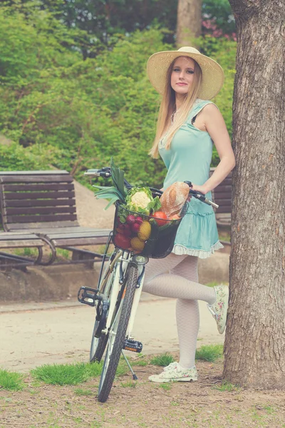 Attractive blonde woman with straw hat standing in the park and posing next to bike with basket full of groceries. — Stock Photo, Image