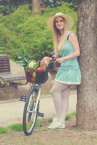 Mulher loira atraente com chapéu de palha em pé no parque e posando ao lado de bicicleta com cesta cheia de compras . — Fotografia de Stock