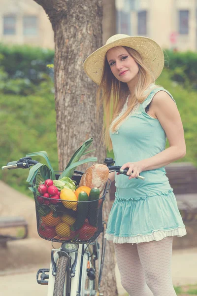 Mulher loira atraente com chapéu de palha em pé no parque e posando ao lado de bicicleta com cesta cheia de compras . — Fotografia de Stock