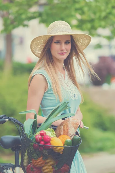 Attraktive blonde Frau mit Strohhut steht im Park und posiert neben Fahrrad mit Korb voller Lebensmittel. — Stockfoto