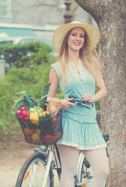 Attractive blonde woman with straw hat standing in the park and posing next to bike with basket full of groceries. — Stock Photo, Image