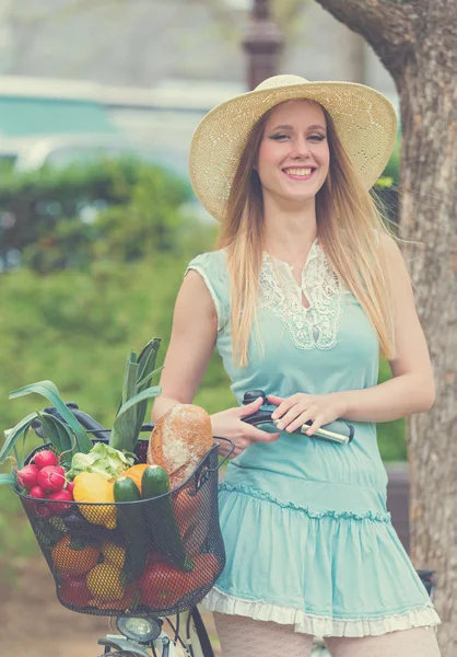 Attraente donna bionda con cappello di paglia in piedi nel parco e in posa accanto alla bici con cesto pieno di generi alimentari . — Foto Stock