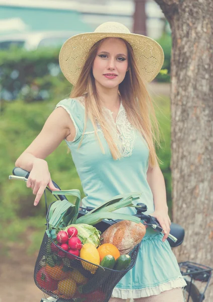 Attractive blonde woman with straw hat standing in the park and posing next to bike with basket full of groceries. — Stock Photo, Image