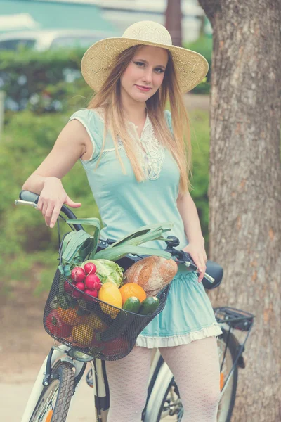 Attraktive blonde Frau mit Strohhut steht im Park und posiert neben Fahrrad mit Korb voller Lebensmittel. — Stockfoto