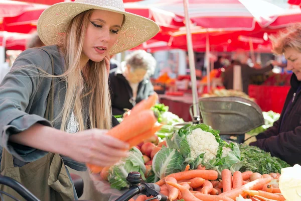 Ελκυστική ξανθιά κοπέλα με ψάθινο καπέλο αγοράζουν λαχανικά στο marketplace. — Φωτογραφία Αρχείου