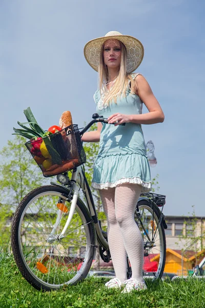 Attractive blonde woman with straw hat posing next to bike with basket full of groceries. — Stock Photo, Image