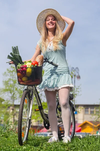 Attractive blonde woman with straw hat posing next to bike with basket full of groceries. — Stock Photo, Image