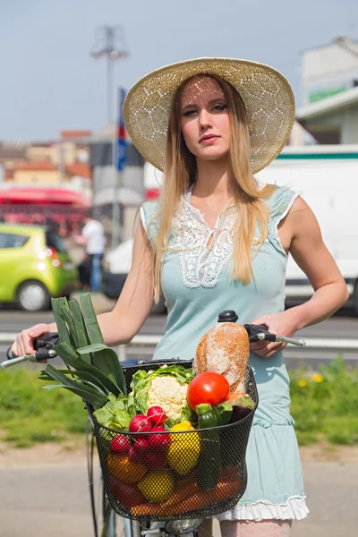 Attractive blonde woman with straw hat posing next to bike with basket full of groceries. — Stock Photo, Image