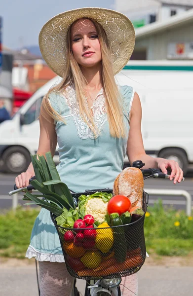 Attractive blonde woman with straw hat posing next to bike with basket full of groceries. — Stock Photo, Image