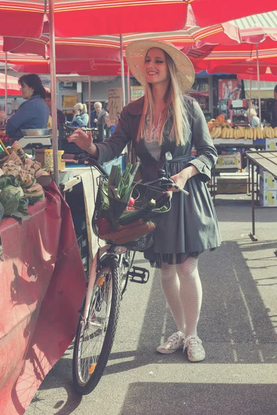 Atractiva mujer rubia con sombrero de paja y bicicleta en el mercado . — Foto de Stock