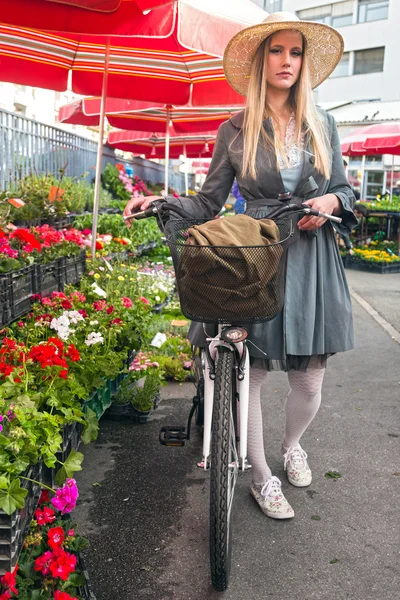 Attractive blonde girl with straw hat and bike on Marketplace. — Stock Photo, Image