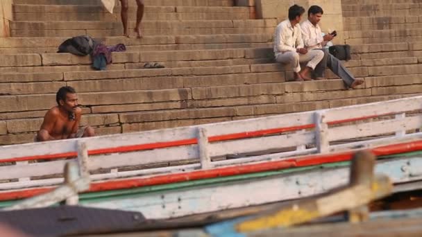Muži sedící u schodiště řeky gangy ve Váránasí, lodě vpředu. — Stock video