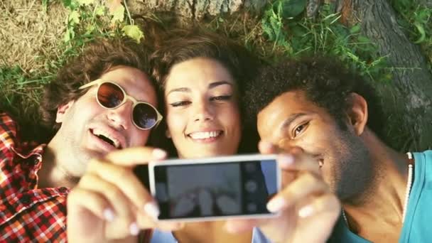 Троє молодих людей беруть селфі, лежачи на траві — стокове відео