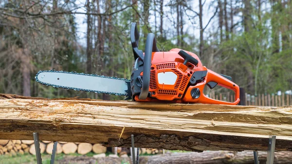 Orange hand gasoline chainsaw on wooden log in metal sawhorse. Professional power saw with black handle and sharp teeth in a rotational chain on guide bar. New cutting tool for wood sawing. Forestry.