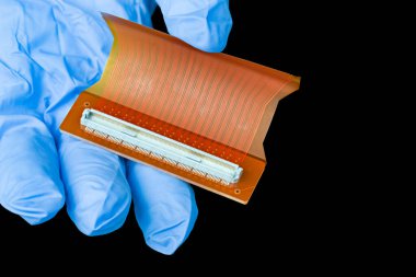 Elektronik esnek devre kartı, siyah arka planda izole edilmiş teknisyen eliyle. Turuncu plastik PCB ve beyaz konnektör tutan mavi eldivenli parmaklar. Veri sinyalleri LCD ekranına iletiliyor.