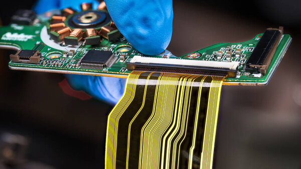 Flat plastic strip flexible cable on printed circuit board on a dark background. Hand in blue glove holding green PCB with integrated motor stator and electronic components as micro chip or resistors.