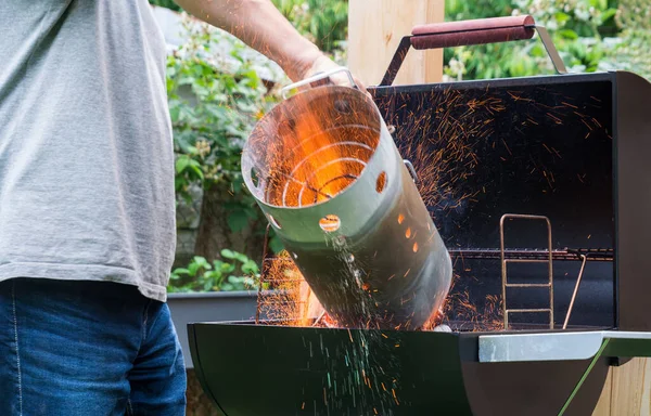 Kömür ya da tütsü tutuşturmak için baca mangalı kullanılacak. Sıcak kıvılcımlar silindirik metal tüp ışık konisiyle havada uçuşuyor. Bahçe barbeküsü için ateş yakan adam.