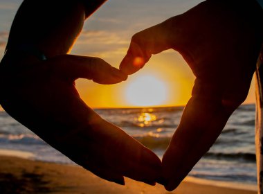Gün batımı ellerden yapılmış bir kalple parlar. Sevgi dolu bir çift elleriyle kalp yaptı ve gün batımını izletti. Deniz ve kumsalda gün batımı.