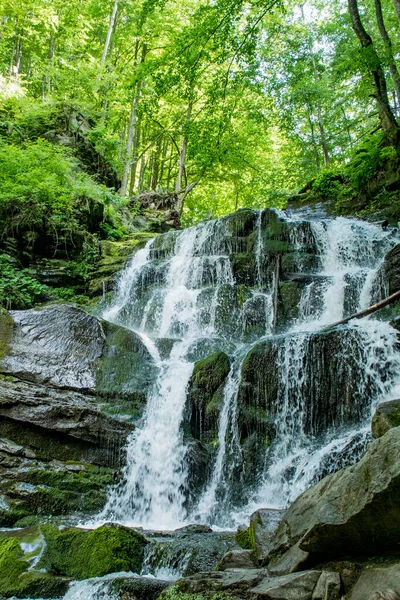 Beautiful and high waterfall in the forest. The waterfall is surrounded by forest, trees, grass. Landscape with a waterfall for wallpaper, magazines, travel.