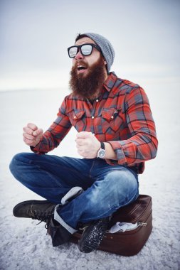hyped up hobo like hipster with manly awesome beard and sunglasses sitting on retro suitcase in empty desolate salt flats clipart