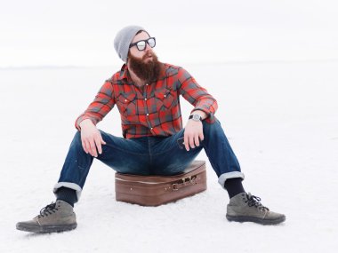 bearded hipster sitting on suitcase in salt flats waiting clipart