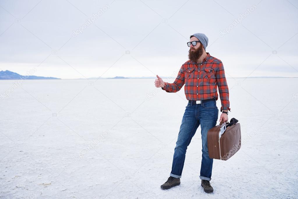 nonchalant entitled bearded hipster hitch hiker thumbing for a ride in empty vast salt flats with retro suitcase