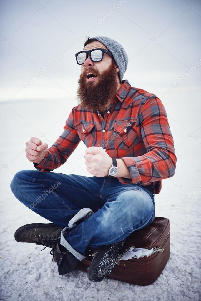 hyped up hobo like hipster with manly awesome beard and sunglasses sitting on retro suitcase in empty desolate salt flats