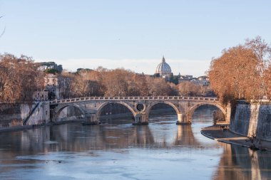Bu görüntü, antik bir taş köprünün altından birden fazla kemerle akan bir nehrin geniş bir görüntüsünü yakalar. Arka planda, Aziz Peter Bazilikası 'na benzeyen kendine özgü kubbeli bir bina var.