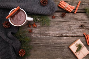 Bir kupa sıcak çikolata, baharatlar, gri örgü eşarp, köknar ağacı dalları, koniler, kırsal ahşap masa arka planında el işi hediyesi. Kış ve Noel konsepti. Baharatlı kakao, yeni yıl, üst görünüm, kopyalama alanı